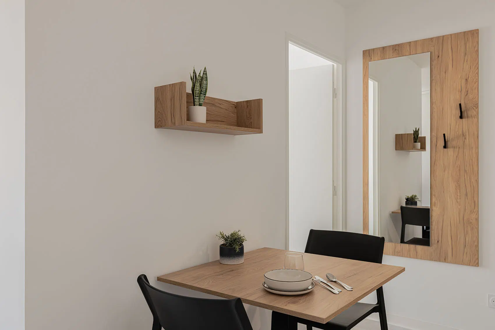 Table de repas finition bois avec chaises noires, étagère murale et grand miroir sur panneau assorti avec patères.