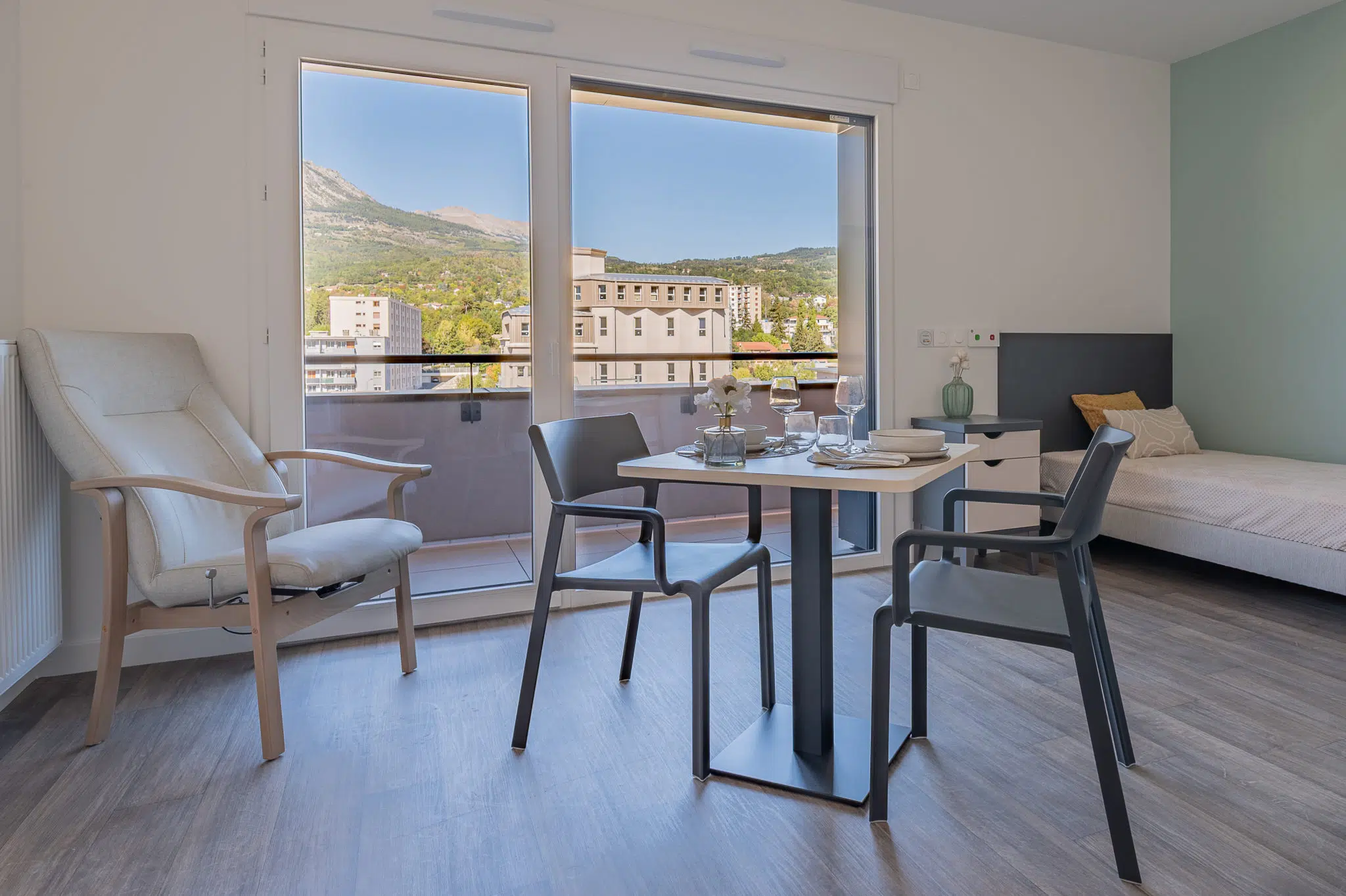 Table, chaises avec accoudoirs et fauteuil devant la baie vitrée d’un appartement de la résidence Le Bocage.