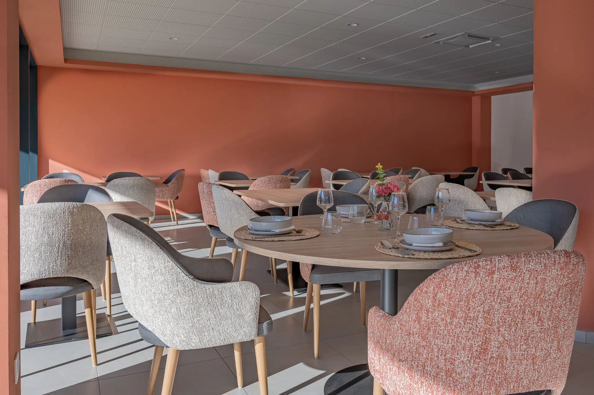 Tables et chaises aux tons terracotta, beige et gris dans une salle de restaurant lumineuse.